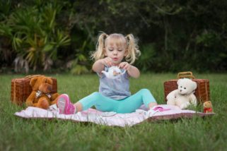 A blonde-haired little girl in pigtails pouring tea for her teddy bear friends at a picnic.